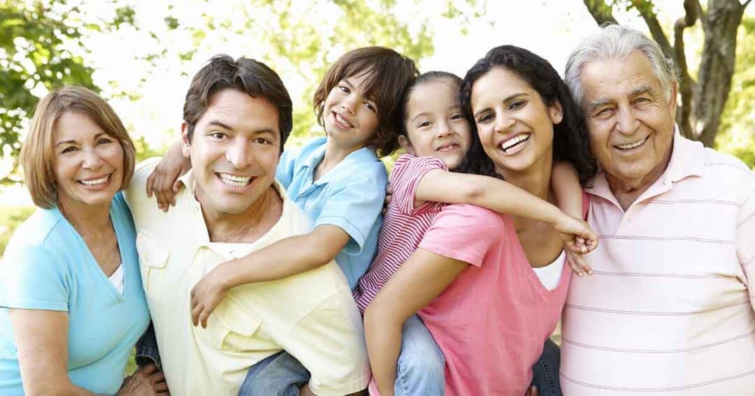 Trusted Messengers 2 A happy family of six stands outdoors, smiling. Two children are being playfully held by a man and woman, surrounded by an older couple. They wear casual summer clothes, and trees are visible in the background.