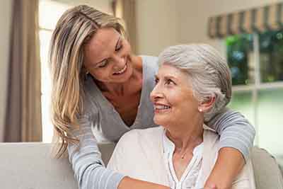 A young woman with long blonde hair smiles while embracing an older woman with short gray hair. They are indoors, sitting on a couch in a cozy room.