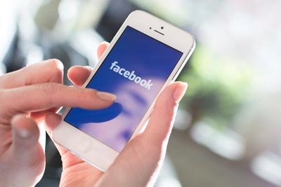 A person holding a smartphone displaying the Facebook app on the screen, with their thumb poised to tap. The background is softly blurred.