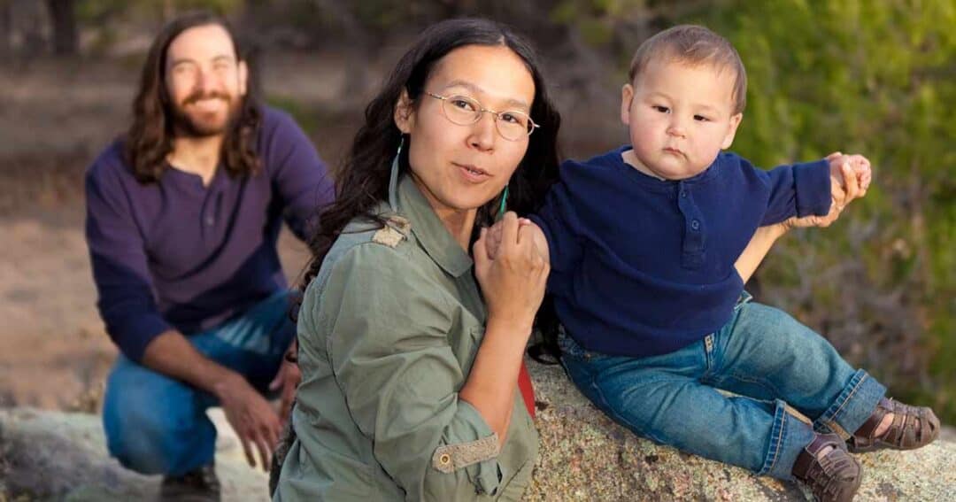 Health Equity in Allergies, Asthma and Immunology 9 A mother holding a baby boy sits on a rock outdoors, looking at the camera. The baby is wearing a blue shirt and jeans. A man with long hair and a beard kneels in the background, smiling. Trees are visible in the background.