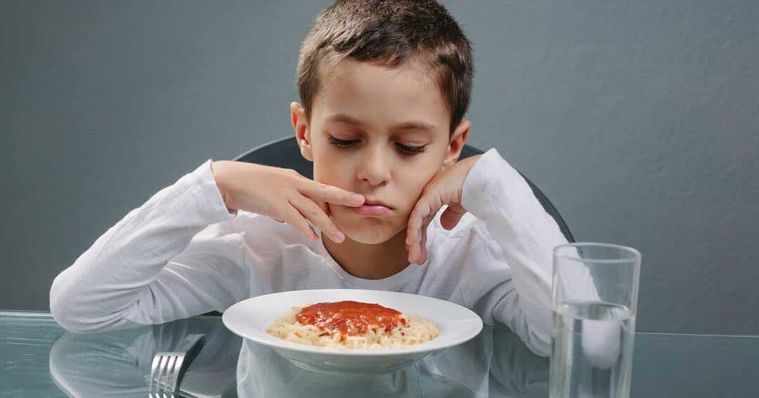 Mental Health and Living with Food Allergies 2 A young boy in a white shirt stares anxiously at a plate of spaghetti with tomato sauce, worried about possible food allergies. He sits at a glass table with a glass of water nearby, set against a plain gray background.