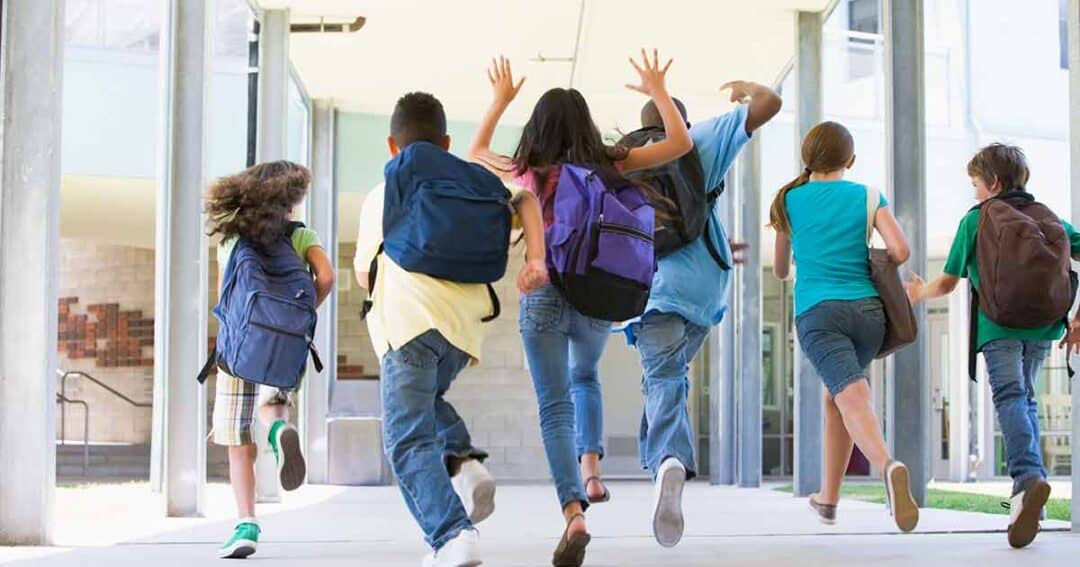 A group of five children with backpacks are joyfully running down a covered walkway, possibly at a school, with their arms raised. They appear to be heading towards a building. The atmosphere is lively and energetic.