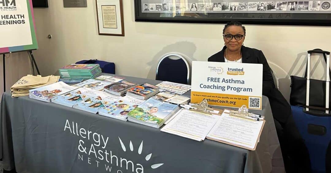 Trusted Messengers 5 Laonis Quinn RN, MSN, AE-C sits at an information booth with a sign that reads "FREE Asthma Coaching Program." The table is covered with brochures, magazines, and clipboards. A banner behind the table proudly displays "Allergy & Asthma Network.