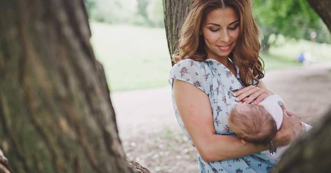 Pregnancy and Allergies 4 A woman with long brown hair wearing a floral blouse is smiling down at a baby she is holding. They are outdoors, surrounded by trees and greenery.