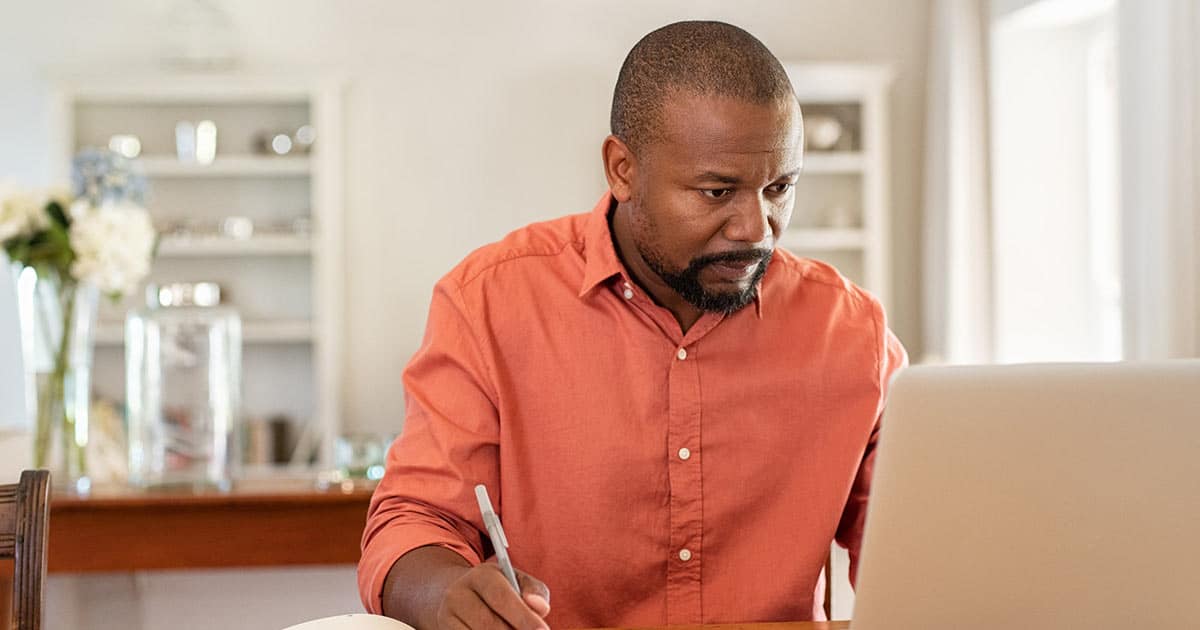 A man in an orange shirt sits at a table, focused on his laptop. He holds a pen in one hand. The background shows a bright room with shelves and a vase of flowers.