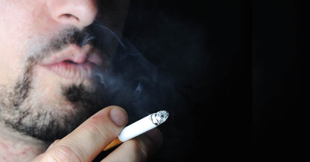 A person with facial hair holds a lit cigarette close to their mouth, exhaling smoke against a dark background. The image focuses on the cigarette and the smoke.