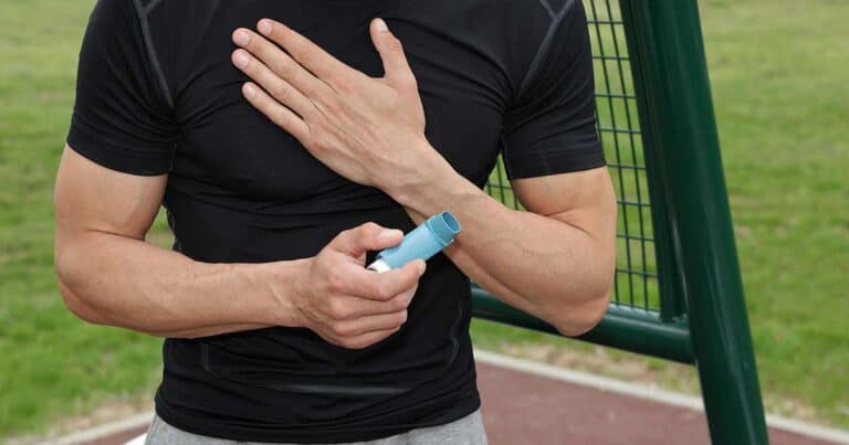 A person in a black shirt holding an inhaler in one hand, with the other hand placed on their chest. They are standing outdoors near a green structure, with blurred grass in the background.