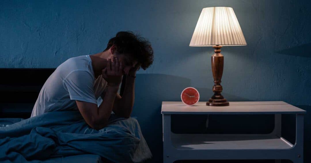 A person sits on a bed in a dimly lit room, head in hands, appearing thoughtful or stressed. A lamp glows on a bedside table next to an alarm clock. Battling nocturnal asthma under the calming yet somber blue walls adds to the night's quiet struggle for breath.