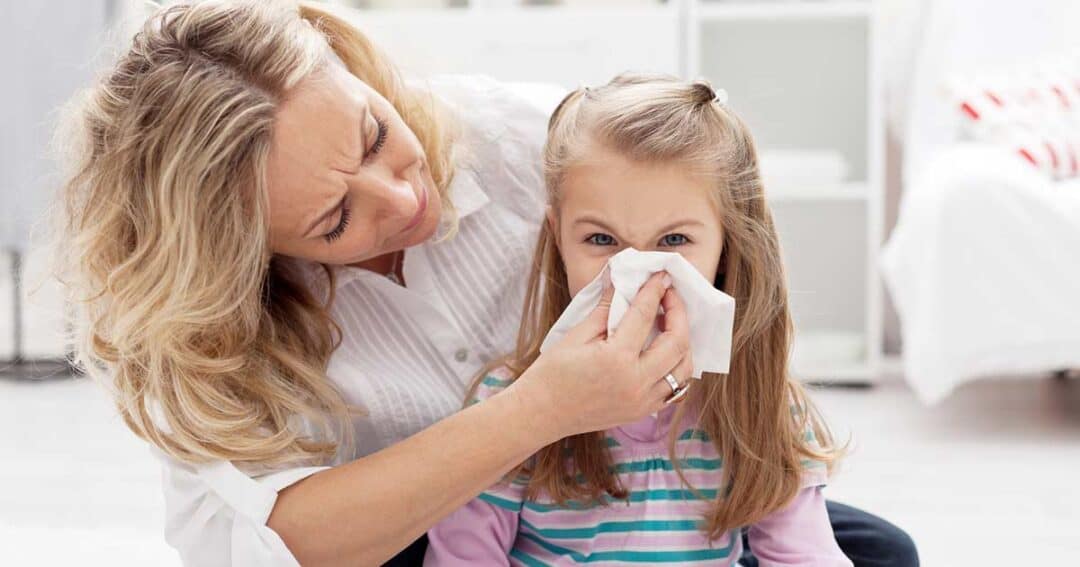 A woman assists a young girl with allergy symptoms, gently helping her blow her nose with a tissue. The girl wears a serious expression, and they are indoors. The softly blurred background reveals a serene, home-like setting.
