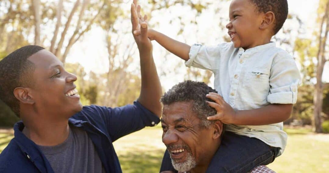 Trusted Messengers 4 Three people outdoors: a child on an older man's shoulders, both smiling. The child is reaching out to high-five another smiling man. They are in a park with trees in the background on a sunny day.