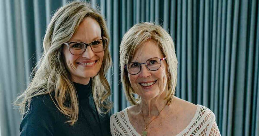Two women with blonde hair and glasses smile at the camera while standing close together in front of grey curtains. One wears a black top; the other wears a white, patterned blouse.