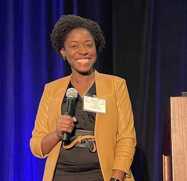 USAsthma Summit 2023: Is Asthma Remission Possible? 2 Woman smiling and holding a microphone, wearing a tan blazer over a black outfit, standing on stage in front of a dark curtain.