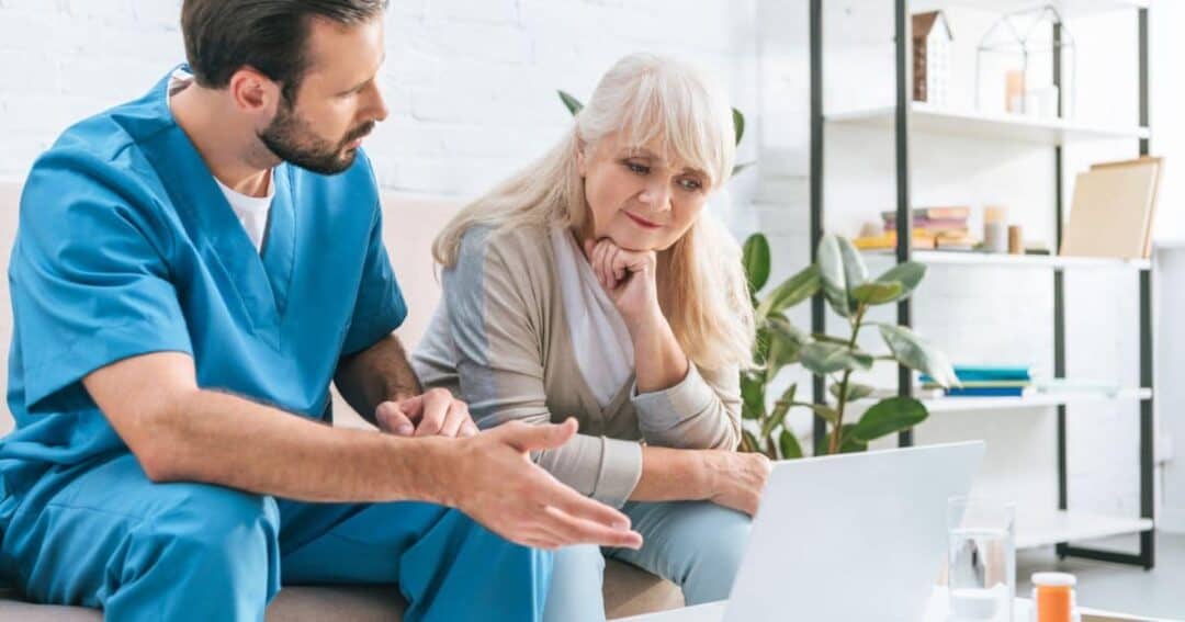 A male nurse in blue scrubs is sitting on a couch and discussing something with an elderly woman with long white hair. They are looking at a laptop screen and he's helping her complete a survey. There is a plant and a shelf in the background.