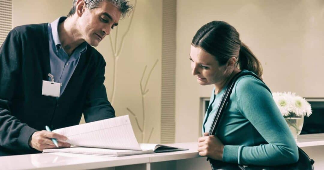 A man in a cardigan and ID badge assists a woman in a teal hoodie at a reception desk. He points at a document while she leans over, observing. A vase with white flowers is on the counter.