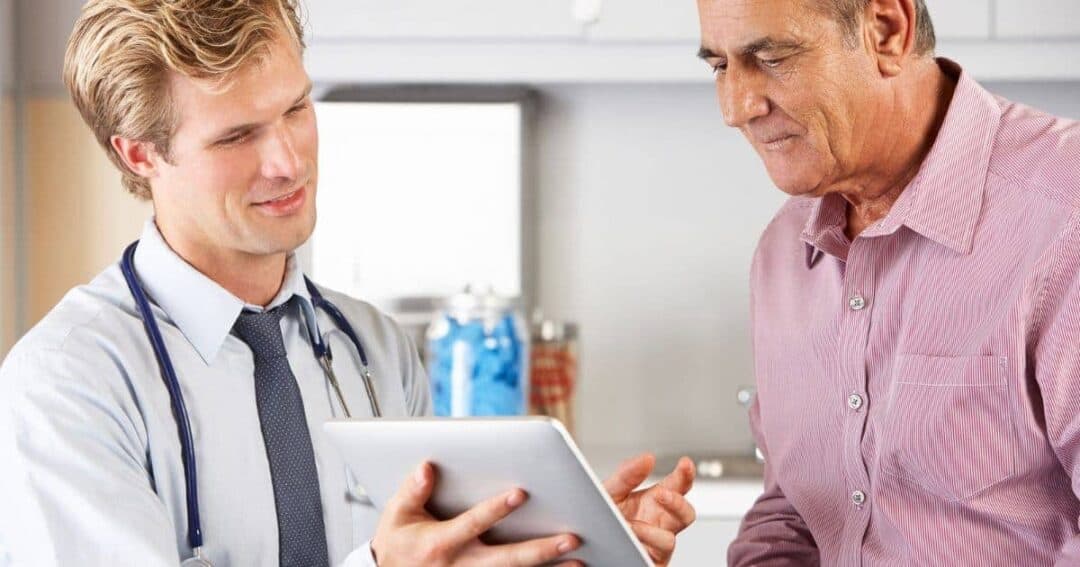 A doctor with a stethoscope around his neck shows a tablet to an older man in a pink shirt. They are in a well-lit room, possibly a clinic, and are engaged in a discussion.