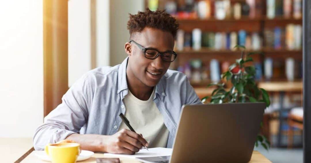 A person wearing glasses sits patiently at a desk, diligently writing in a notebook while looking at a laptop. There's a cup of coffee and a smartphone on the table, suggesting they're deep into their research. The background is adorned with bookshelves and a potted plant.