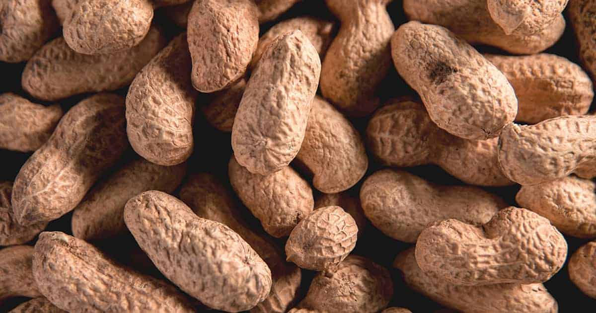 A close-up of several unshelled peanuts grouped together. The textured brown shells are visible, showcasing their organic, rough surface. The image highlights the natural appearance of the peanuts in detail.