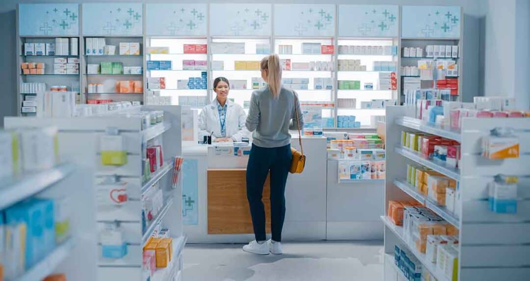 What if I Can’t Afford My Allergy Medications? 2 A woman stands at the counter of a pharmacy, speaking with a pharmacist. Shelves filled with various medications and health products are visible in the background. The space is brightly lit and organized.