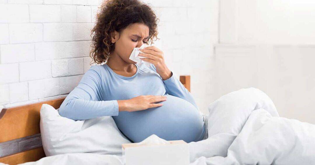 Pregnancy and Allergies 2 Pregnant woman sitting in bed, wearing a blue shirt, blowing her nose with a tissue, and touching her stomach. She looks unwell, with a tissue box nearby. The bed has white sheets and a light, airy atmosphere.