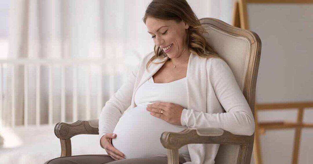 A pregnant woman sits in a cushioned chair, gently cradling her belly with both hands. She is smiling and looking down, wearing a white top and a light-colored cardigan. The background is softly lit and blurred, suggesting a calm, serene setting.