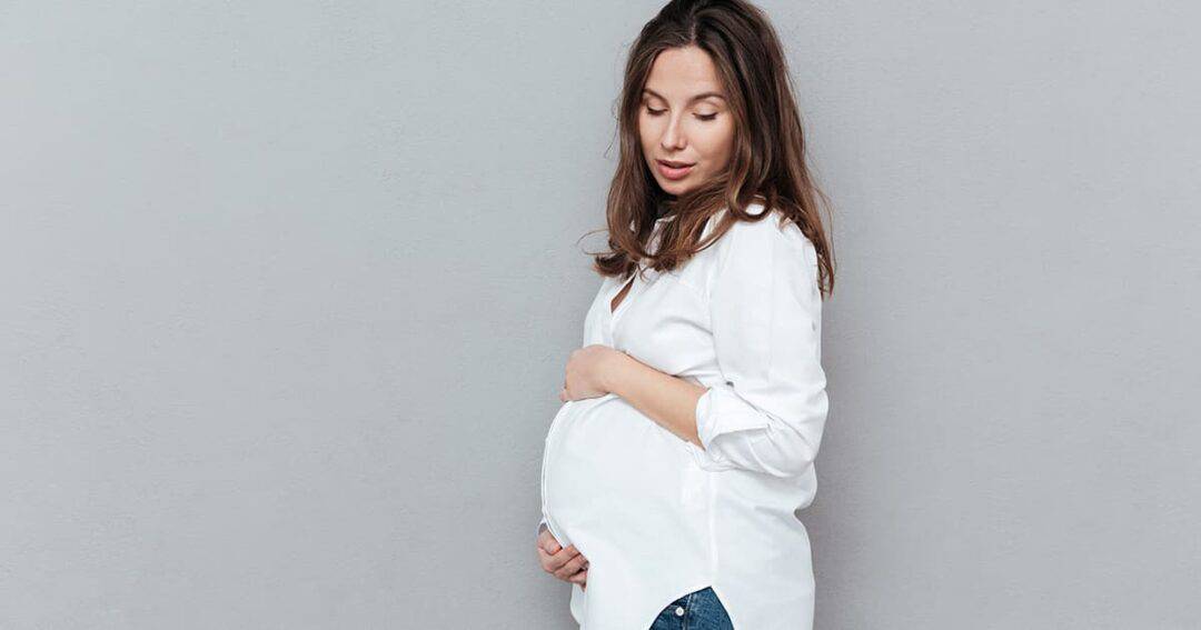 A pregnant woman stands sideways against a gray background, gently holding her belly. She is wearing a white shirt and blue jeans, looking down with a serene expression.