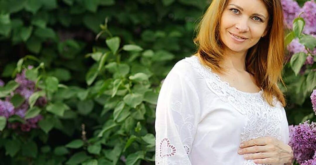 Pregnancy and Allergies 1 A woman with long brown hair is wearing a white dress and standing in front of a lush green background with purple flowers. She is smiling and gently resting one hand on her abdomen.