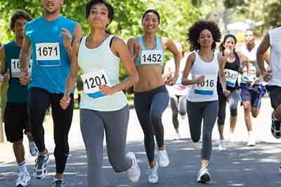 A group of people running in a race, wearing numbered bibs. They are outdoors on a sunny day, surrounded by greenery. The runners appear focused and determined.