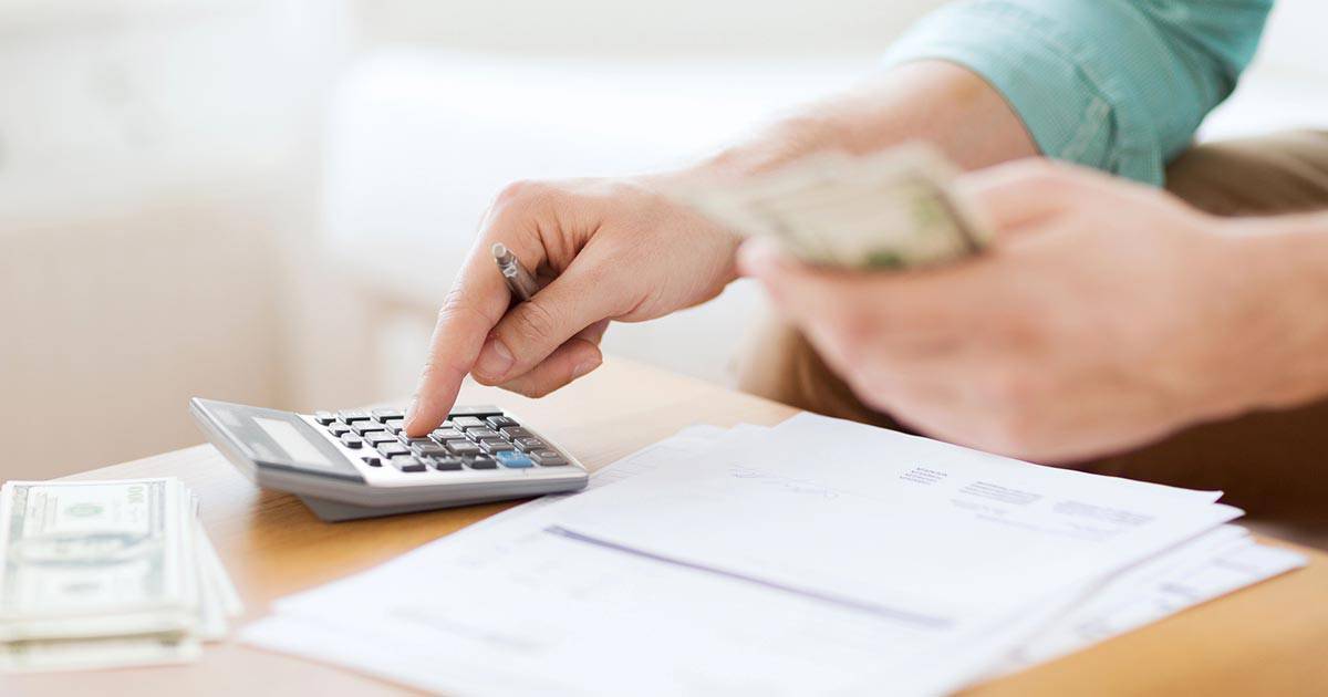A person calculating expenses with a calculator and holding cash. There are papers and dollar bills on the table, indicating financial planning or budgeting activities.