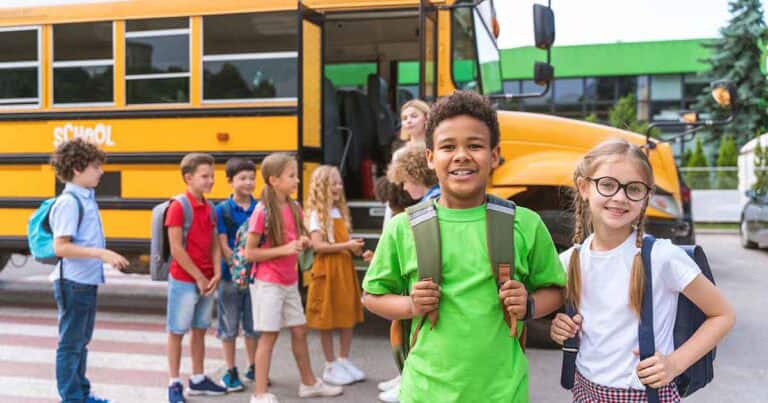 A group of smiling children with backpacks stand near a yellow school bus. Two kids, a boy in a green shirt and a girl in glasses and pigtails, stand in the foreground, while others chat and wait by the bus.