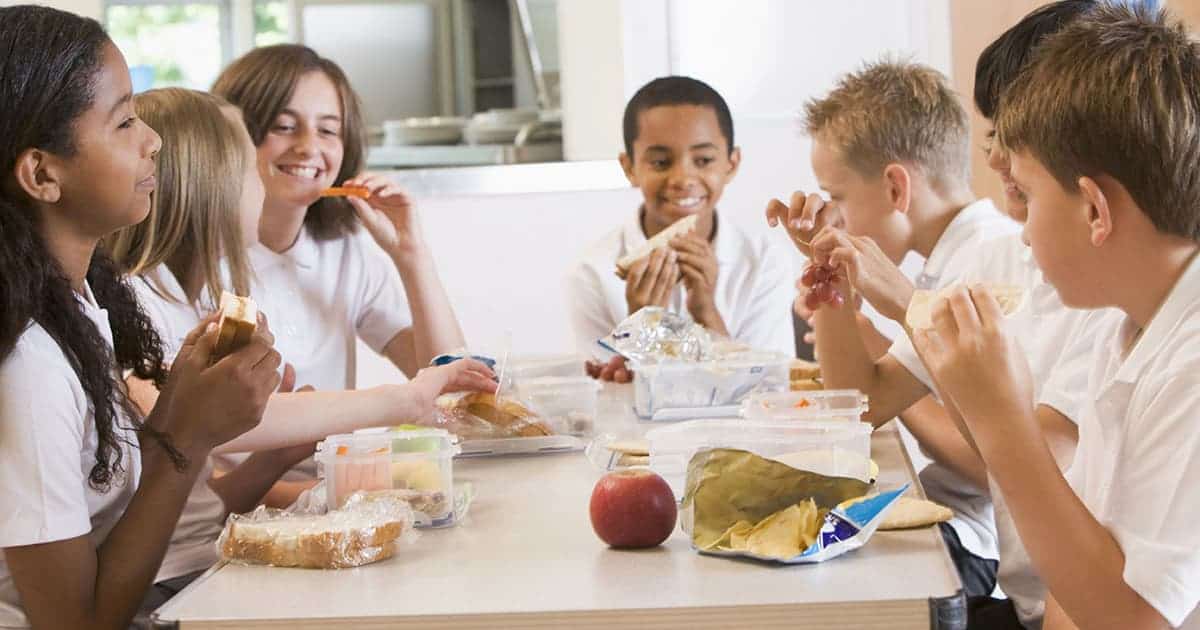 A group of schoolchildren in uniforms sit around a table, enjoying lunch together. They have sandwiches, snacks, and an apple. The atmosphere is cheerful and friendly.