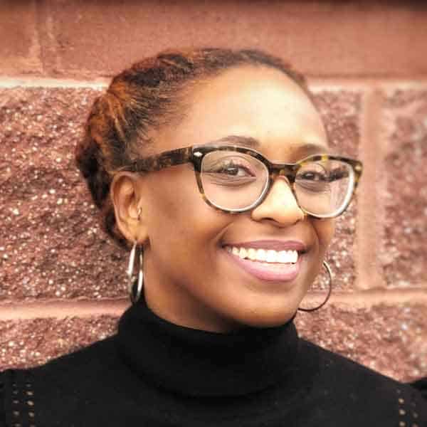 Smiling person with tortoiseshell glasses and hoop earrings stands in front of a brick wall, wearing a black turtleneck. Hair is styled in an updo.