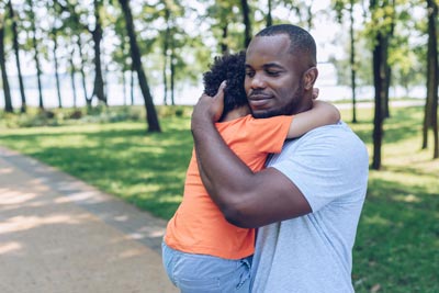 A man gently holds a small child in his arms while standing on a park pathway. The child wears an orange shirt, and they are surrounded by green trees and grass, creating a serene and comforting atmosphere.