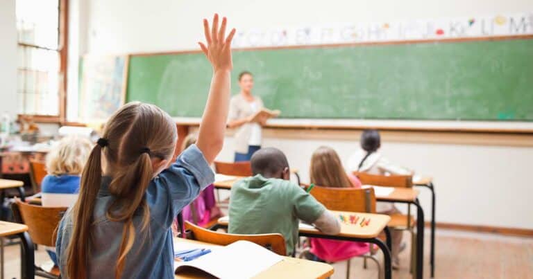 A classroom scene with a young student raising their hand. Several children sit at desks facing a chalkboard, where a teacher stands reading from a book. The classroom is bright and colorful with educational materials on the walls.
