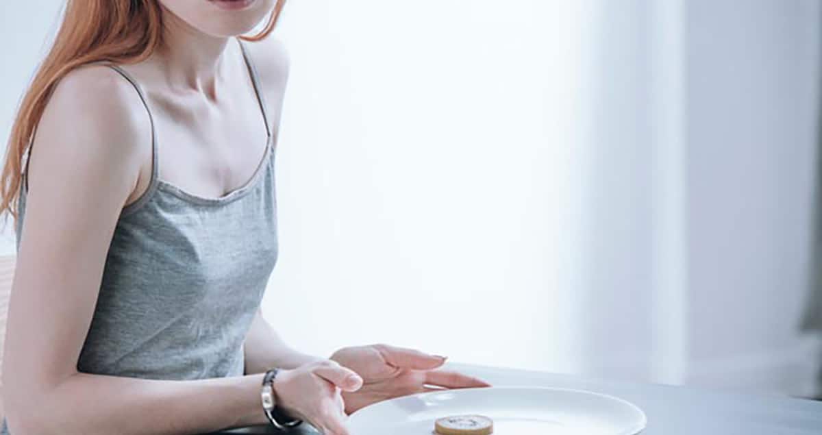 Mental Health and Living with Food Allergies 3 A person with long red hair, wearing a gray tank top, is sitting at a table. They have an empty white plate in front of them with a small pastry on it, reflecting their food anxiety. The background is softly lit with a white curtain.