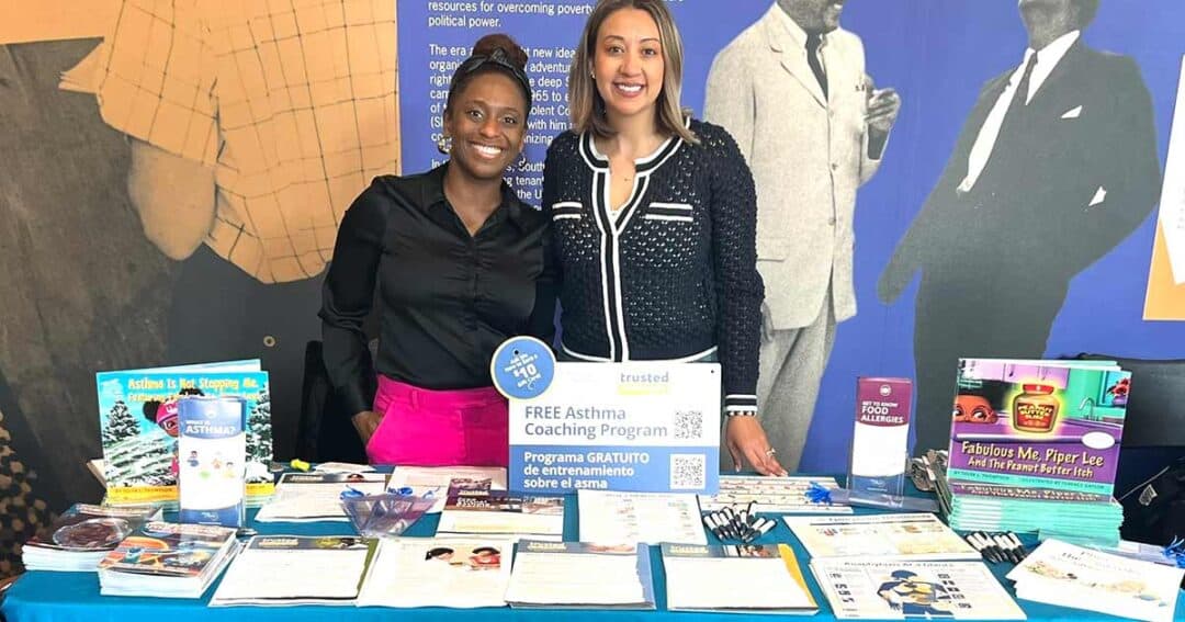 Trusted Messengers 7 Two women stand behind a table with pamphlets and booklets about asthma coaching and health resources. A sign reads “Free Asthma Coaching Program.” The background features images of historical figures.