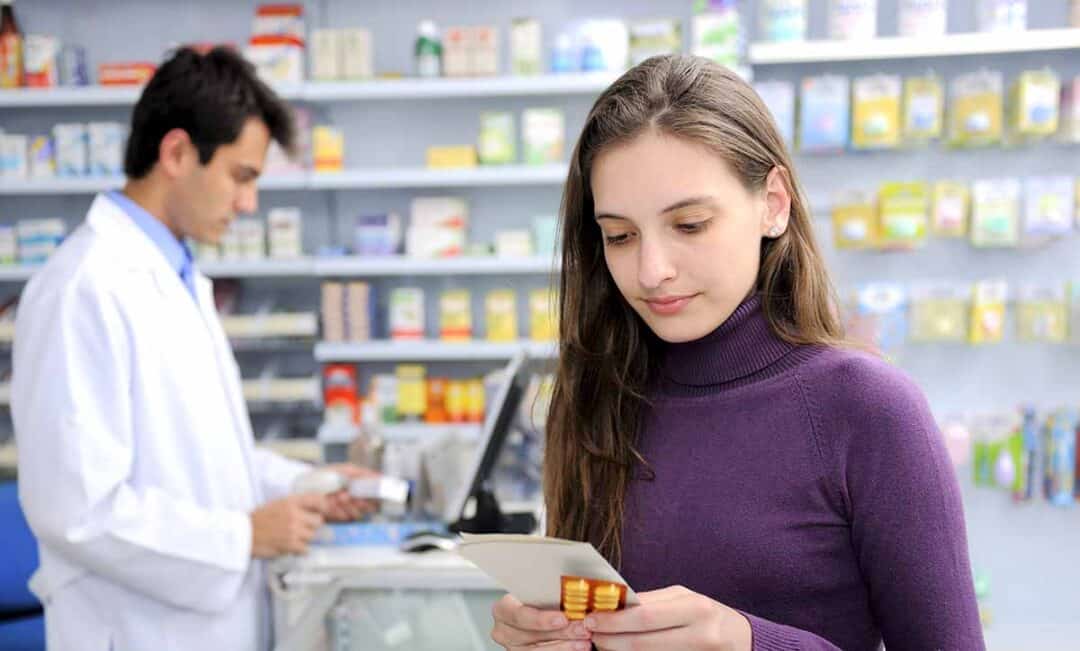 A young woman in a purple sweater examines medication packaging in a pharmacy. Behind her, a pharmacist in a white coat checks prescriptions at the counter. Shelves with various products are visible in the background.