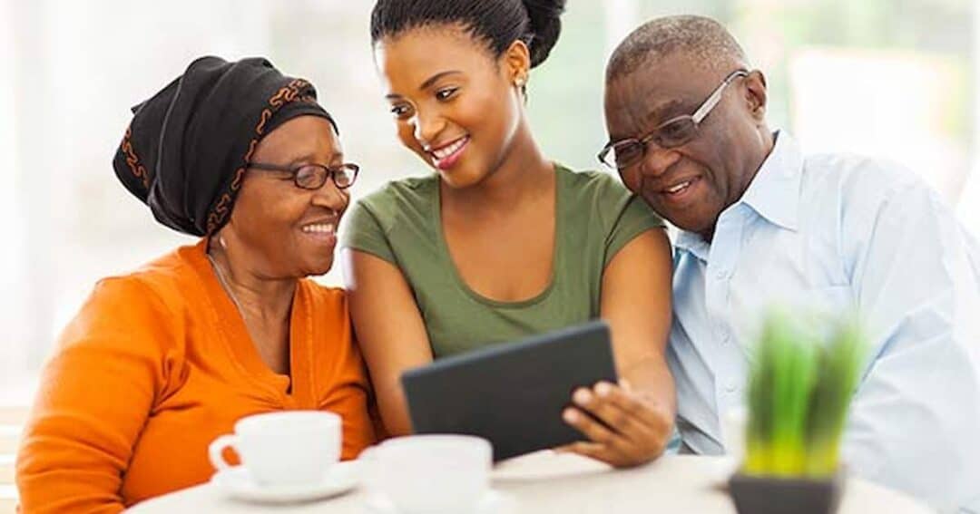 Health Equity in Allergies, Asthma and Immunology 12 A young woman sits between an elderly woman in an orange cardigan and an elderly man in glasses. They are looking at a tablet while sitting at a table with cups and a small plant. All are smiling and appear engaged with the device.