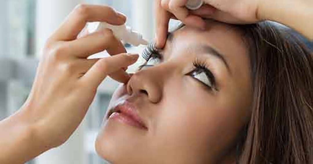 Person tilting their head back, applying eye drops to their right eye. Close-up shot focusing on their face, with hands gently holding the dropper above the eye. The background is softly blurred.