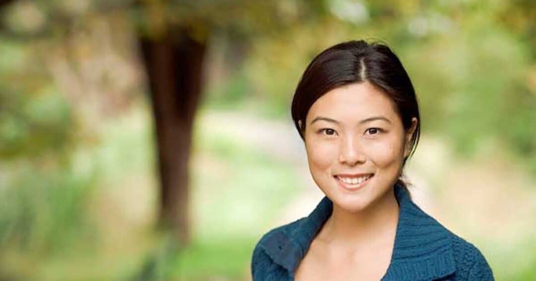 A person with long hair smiles at the camera while standing outdoors. The background is a blurry, lush scene with green trees and grass. The individual is wearing a blue top.