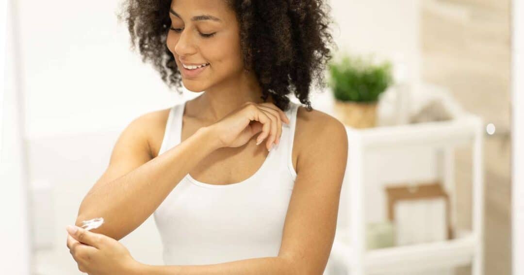 What is Moderate to Severe Eczema? 8 A person in a white tank top is applying eczema treatment lotion to their elbow in a bright bathroom. The background includes shelves with plants and towels. They are smiling and looking at their arm.