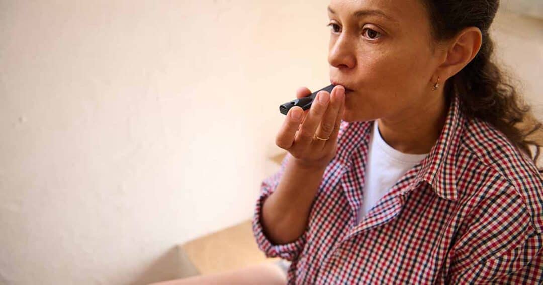 A person wearing a red and white checkered shirt holds a vape pen to their mouth, appearing thoughtful. The background is a neutral, light-colored wall.