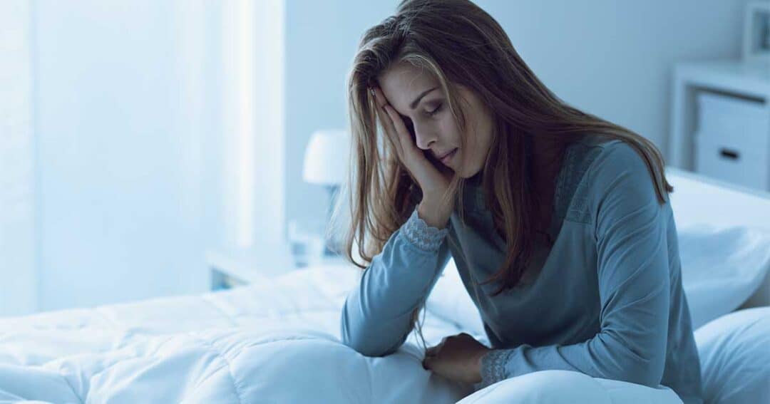 In the blue-lit room, a woman sits on her bed, resting her head with a tired expression. Wearing a long-sleeved top, she appears to be exhausted from lack of sleep because of night time asthma.