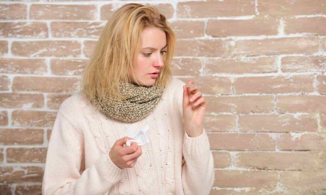 A woman with blonde hair holds a tissue and medicine bottle, suggesting she might be battling allergies. Standing in front of a brick wall, she wears a knitted scarf and light sweater, peering at the bottle with a concerned expression.