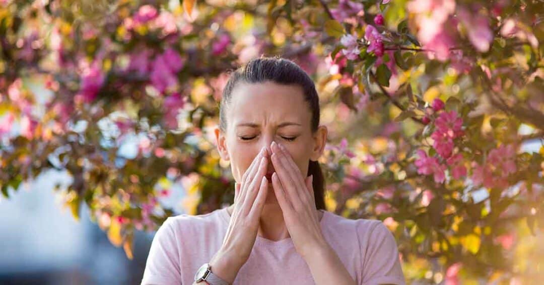 A woman sneezes into her hands, standing outdoors in front of a blossoming tree with pink flowers. She is wearing a light-colored shirt and has her hair tied back.