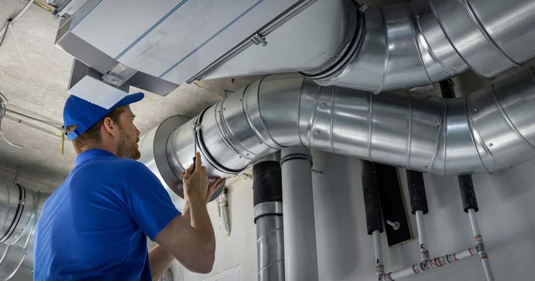 A maintenance worker in a blue uniform and cap inspects large metal air ducts overhead with a flashlight. The ducts are part of an industrial ventilation system in a utility room.