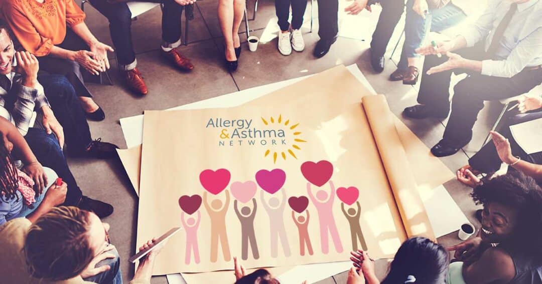 A diverse group of people sit in a circle around a large poster on the floor. The poster shows an illustration of people holding heart shapes, with the text "Allergy & Asthma Network" and a sun logo.